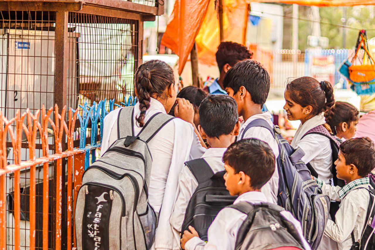Children in uniforms with backpacks standing by a fence outdoors.